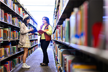 two students standing in an aisle in the library conversing